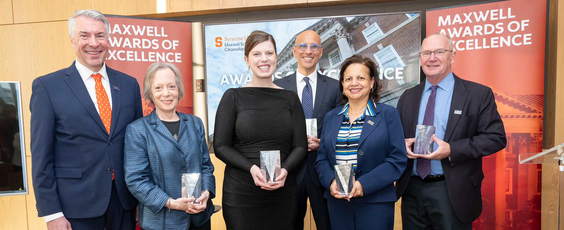 A group of six people stand together holding awards at the Maxwell Awards of Excellence event. They are dressed in formal attire, in front of a backdrop displaying the event's name.