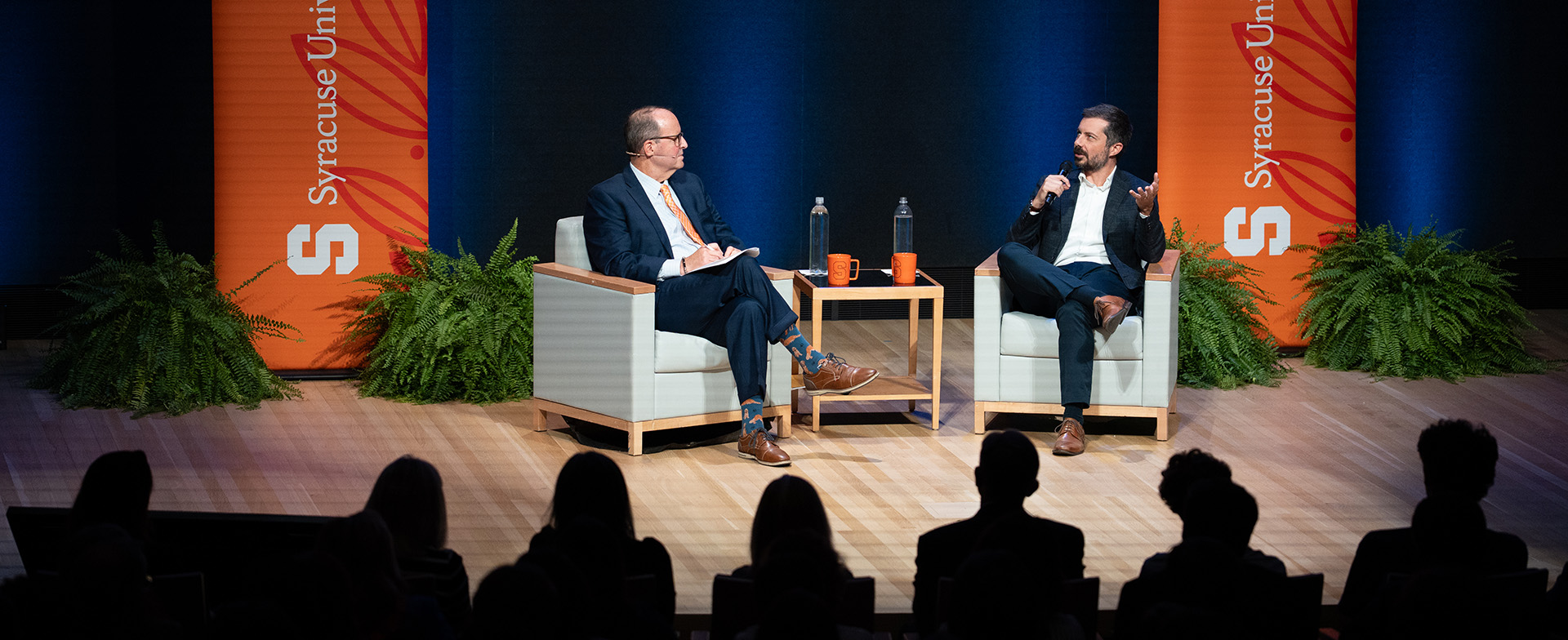 Two people sitting on stage in a discussion at a Syracuse University event, with audience in the foreground and a backdrop featuring the university's logo.