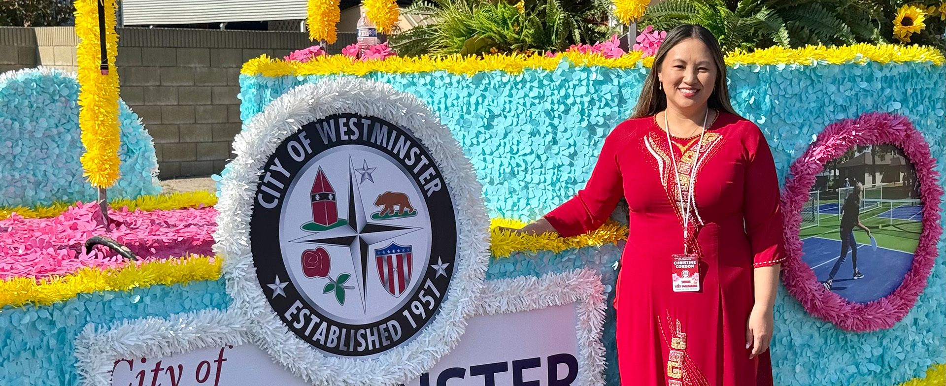 A person stands smiling next to a decorative float with vibrant pink, blue, and yellow floral arrangements. The float features the City of Westminster seal and text indicating "Established 1957."