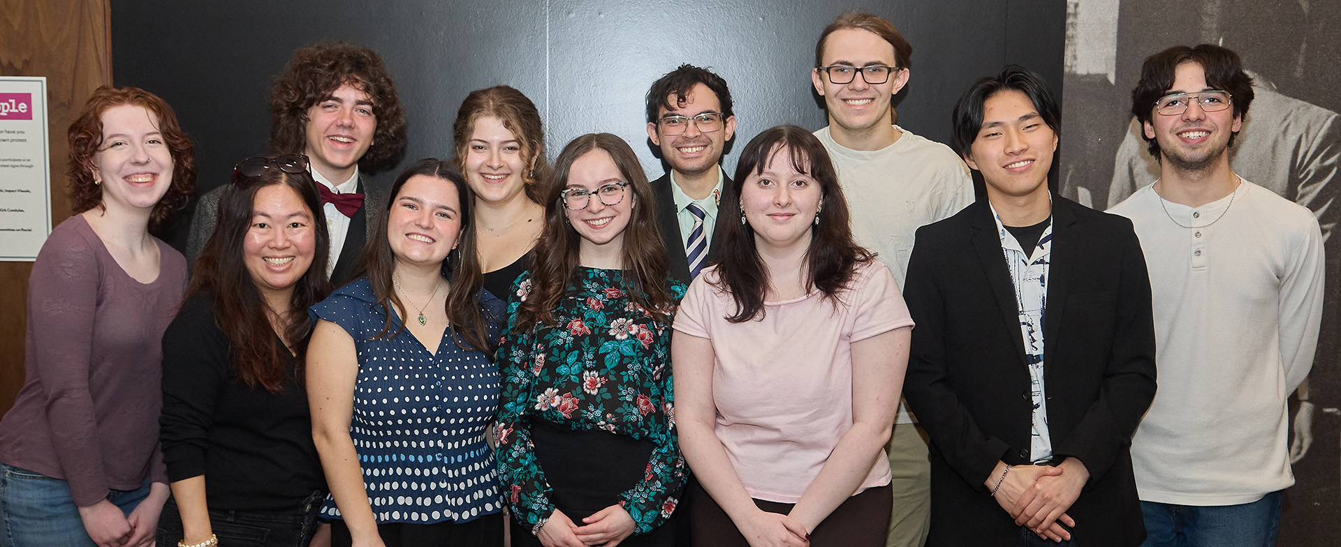 A group of eleven people poses together and smiles for a photo in front of a dark wall with a partial historical mural or exhibit display visible in the background. The people are dressed in a mix of casual and semi-formal attire.