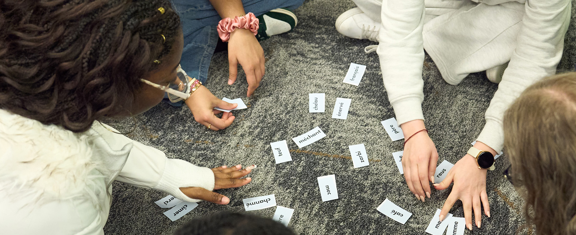 A group of individuals sitting on the floor, actively sorting and arranging small cards with various words printed on them. The floor has a textured, patterned carpet.