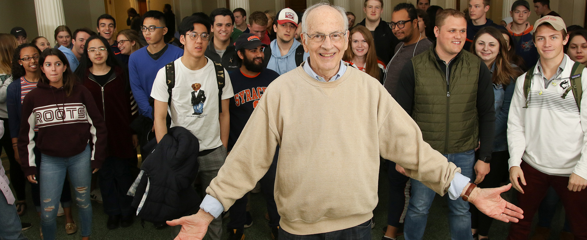 A smiling person stands at the front of a group of diverse students. They are in a casual setting that appears to be indoors. Some students are wearing clothing with various university logos.