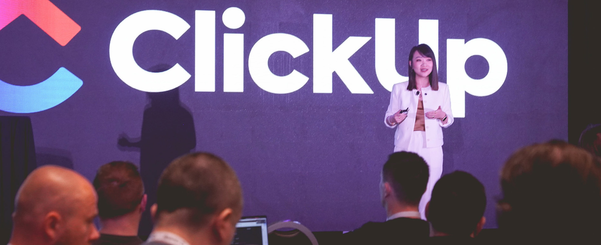 A personspeaks on stage in front of a large illuminated ClickUp logo and branding backdrop, addressing a seated audience at a company or industry event.