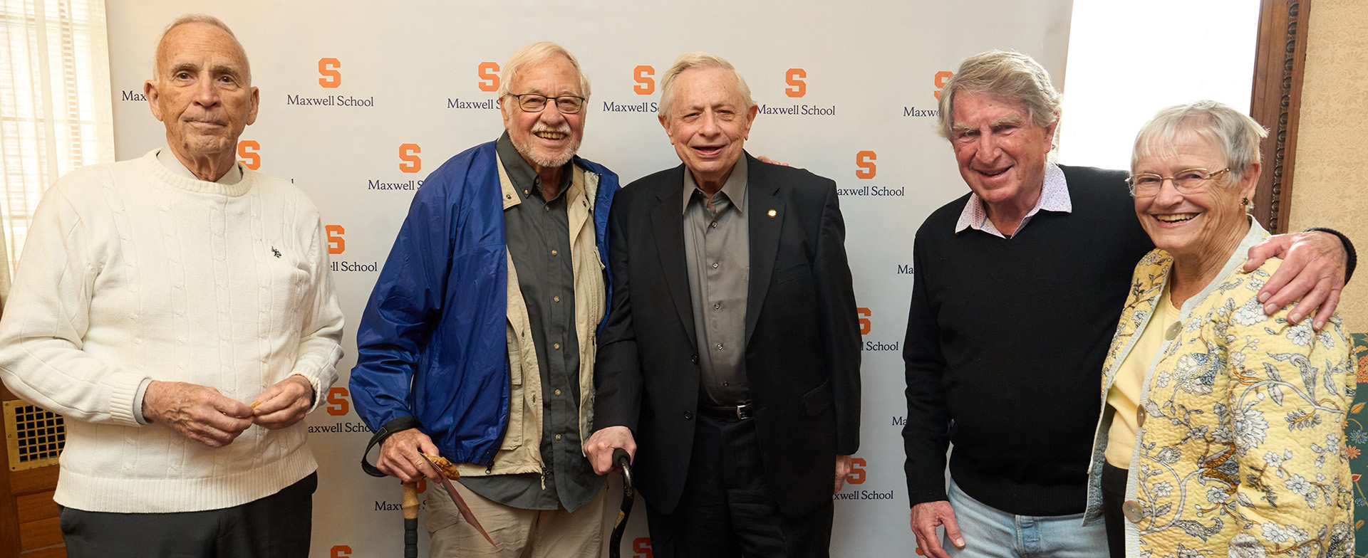 Group of five elderly individuals smiling at the camera, standing in front of a backdrop with the Syracuse University Maxwell School logo.