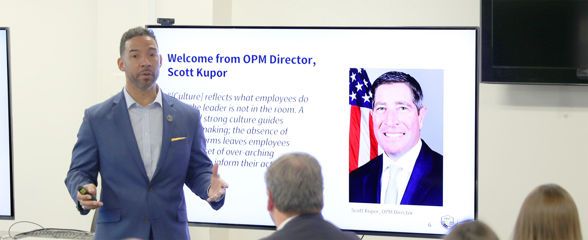 A person is speaking in front of a large screen displaying a presentation. The screen shows a slide titled "Welcome from OPM Director, Scott Kupor" with a photo of the director next to an American flag. Attendees are seated and listening.