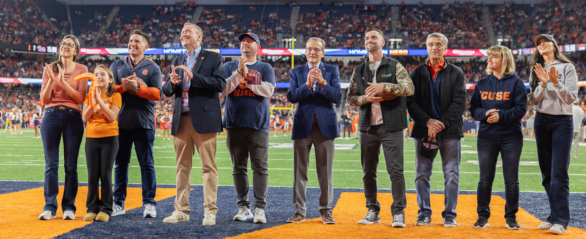 Group of individuals standing on a football field, clapping, at a Syracuse University event.