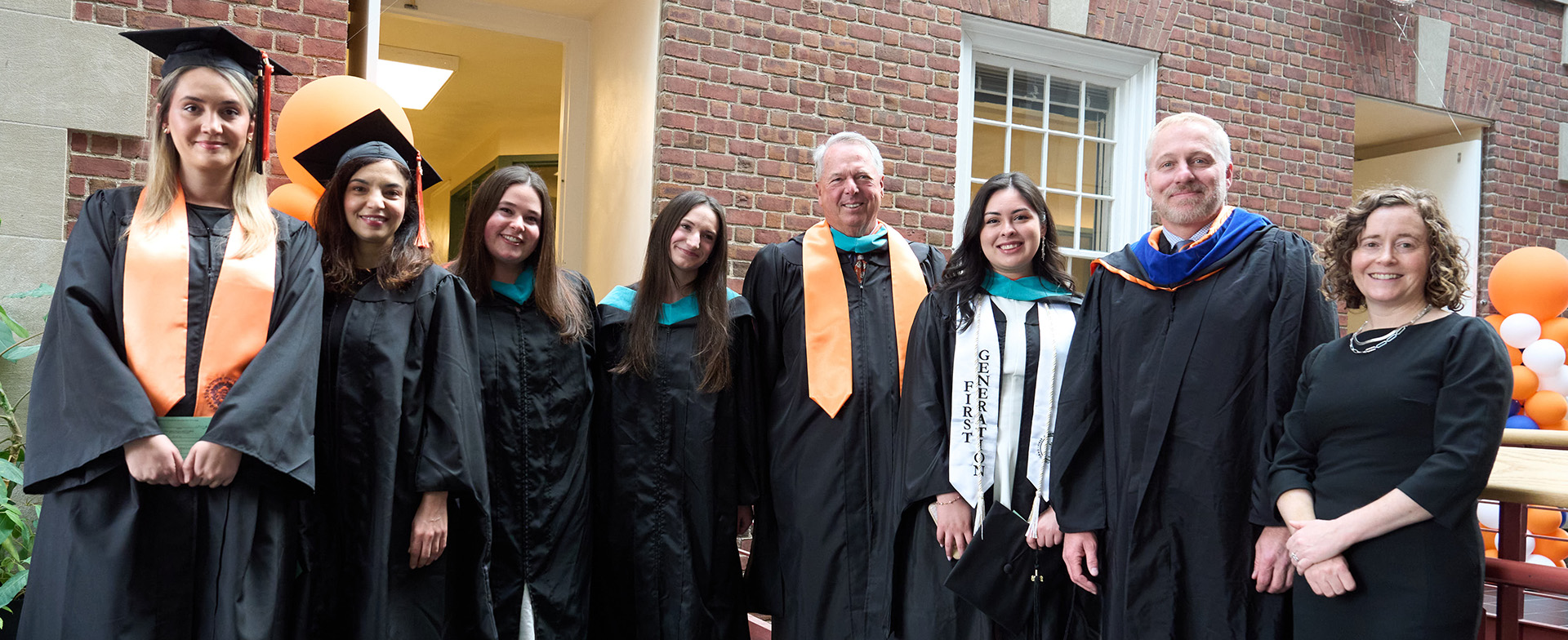 A group of eight people in graduation attire stand together in front of a brick building. Some are wearing mortarboards and stoles with colorful trims. There are orange and blue balloon decorations nearby, suggesting a festive graduation event.
