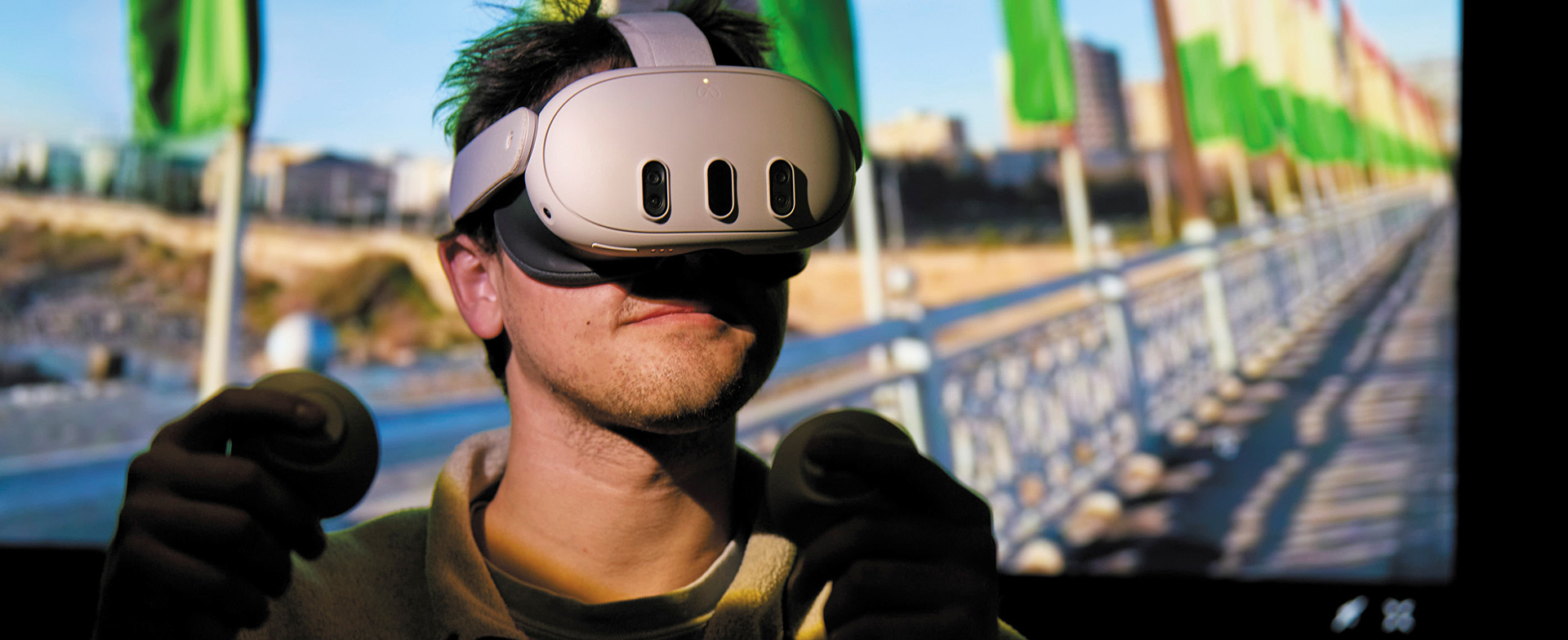 Person using a virtual reality headset and handheld controllers, standing in front of a screen with a row of green flags and a waterfront.