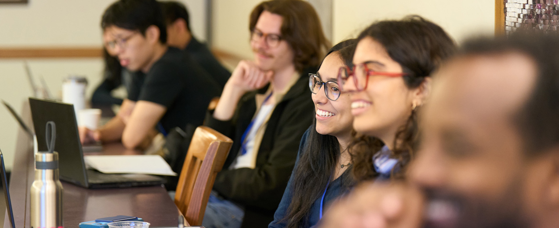 A row of seminar participants seated at tables with laptops, notebooks, and water bottles, smiling and engaged in discussion.