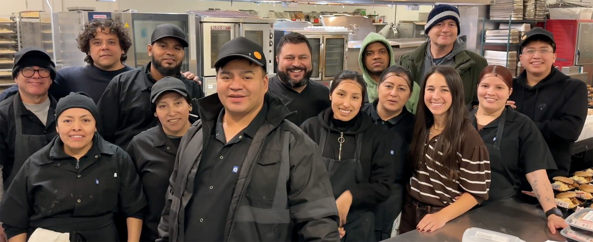 A group of people in a commercial kitchen, all wearing black uniforms and smiling at the camera. They are surrounded by kitchen equipment and trays of food.