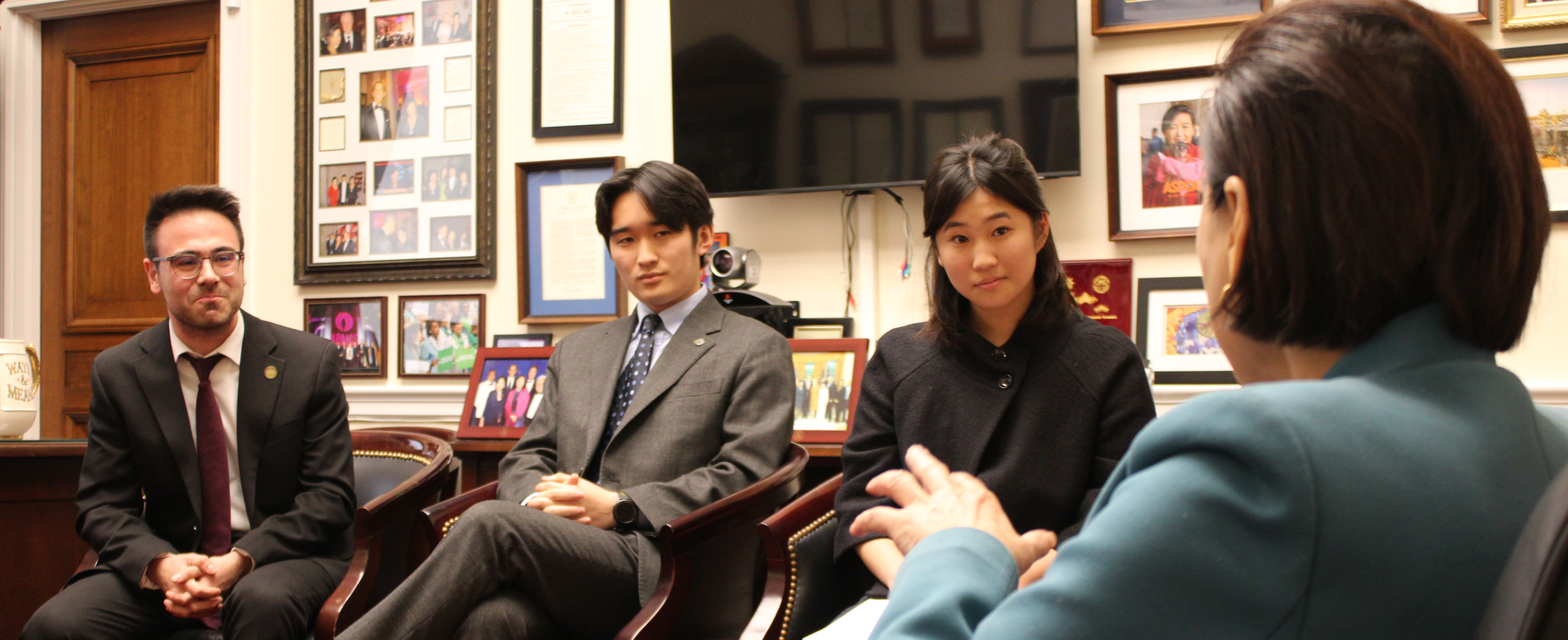 A group of four people is engaged in a discussion in an office setting. The walls feature numerous framed photographs and documents. One person is seated with crossed arms, wearing a suit. Another is dressed in professional attire.