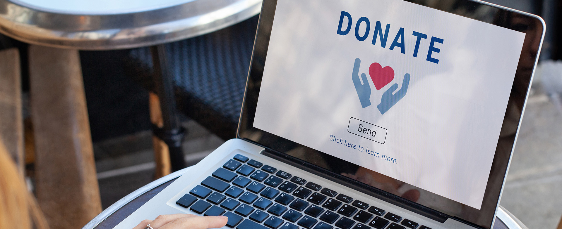 A person seated at a round table is using a laptop. The screen displays the word "DONATE" above an illustration of hands holding a heart, with a "Send" button below.