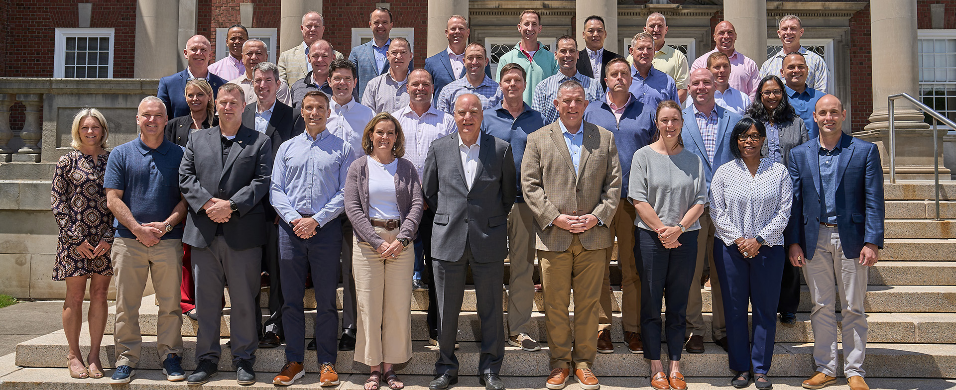 A group of individuals, dressed in business casual attire, stands on the steps in front of a building with columns. They are posed in rows, smiling at the camera.