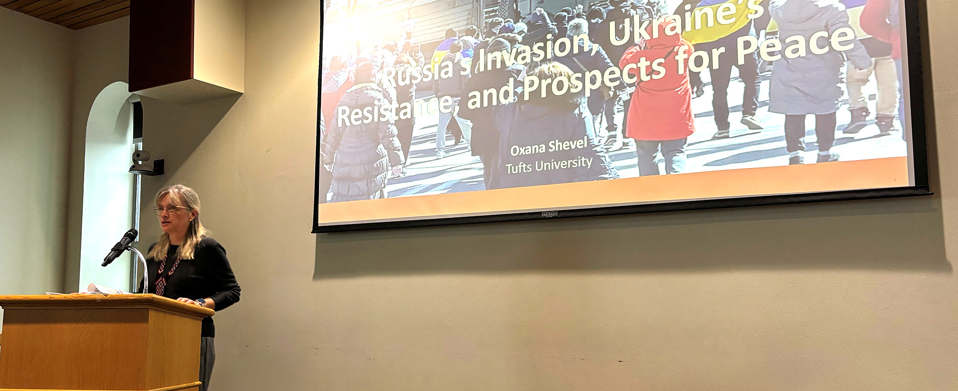 A person stands at a podium speaking in a room. A large screen displays a presentation titled "Russia's Invasion, Ukraine's Resistance, and Prospects for Peace" by Oxana Shevel from Tufts University.