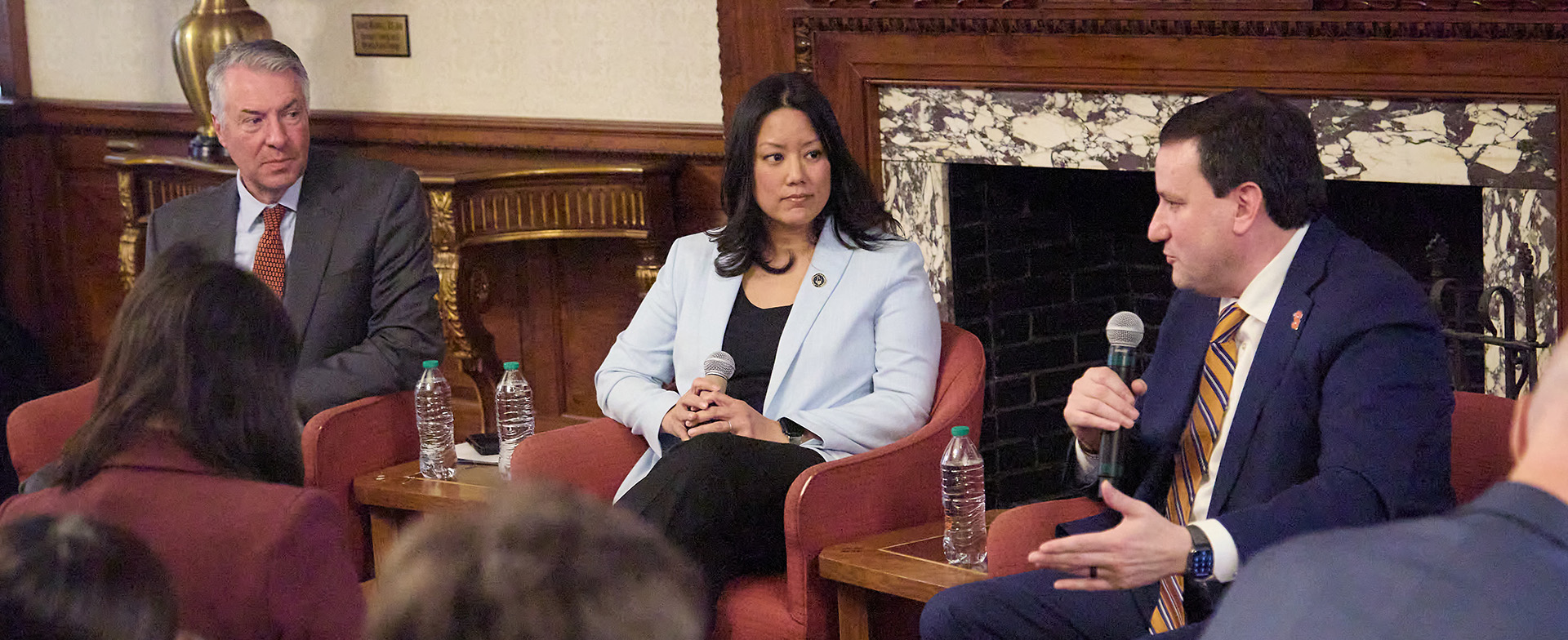 A panel of three individuals is seated on stage participating in a discussion. One person is speaking into a microphone, while the others listen attentively. The setting appears to be a formal room with ornate woodwork and a fireplace.