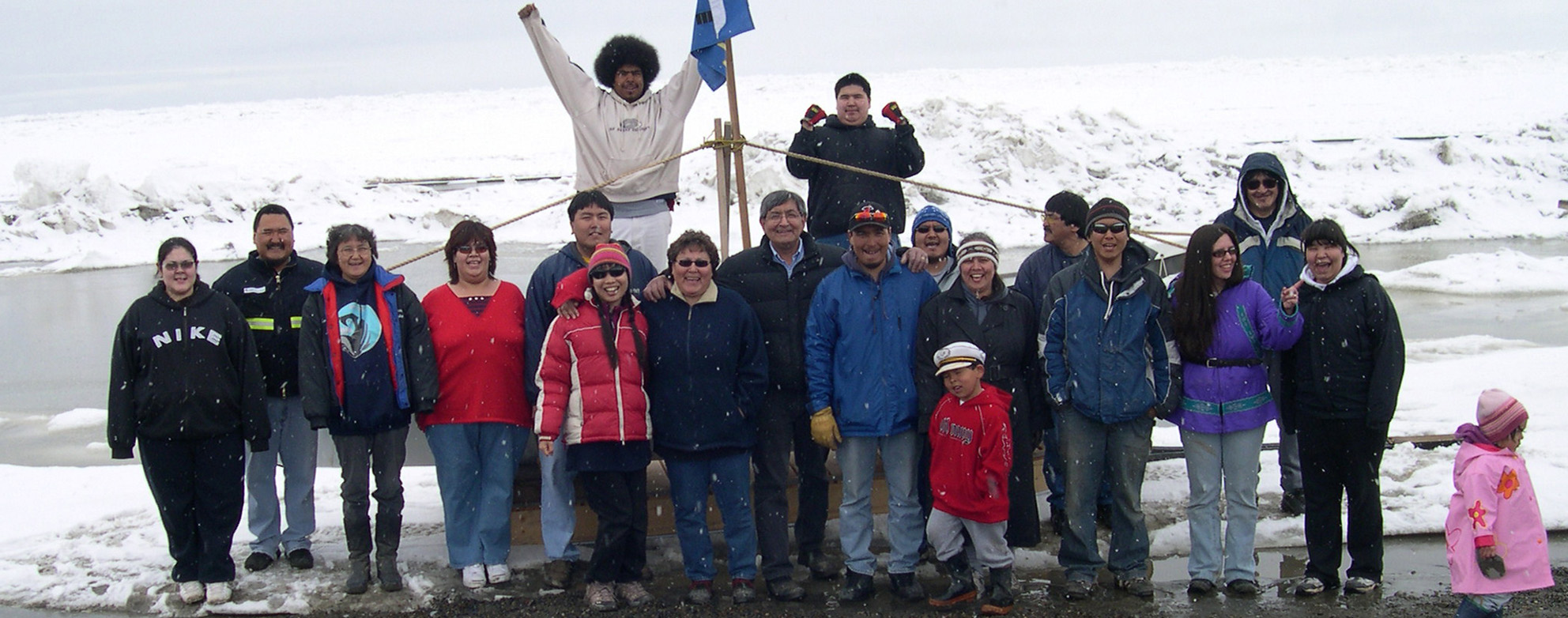 A group of people standing in snowy surroundings near a wooden structure with a flag waving above. Some are wearing winter clothing such as jackets and hats. The background shows snow-covered ground and icy water.