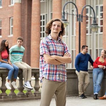 Male student with arms crossed stands in front of Maxwell School, with other students in the background.