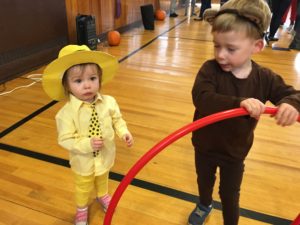 Two brothers dressed as the chracters from curious george. The smaller child wears bright yellow pants and yellow button down, with a pok-a-dot tie and a bright yellow fedora. His older brother stands a few steps way wearing brown jumpsuit and monkey ears while playing with a red hula hoop. 