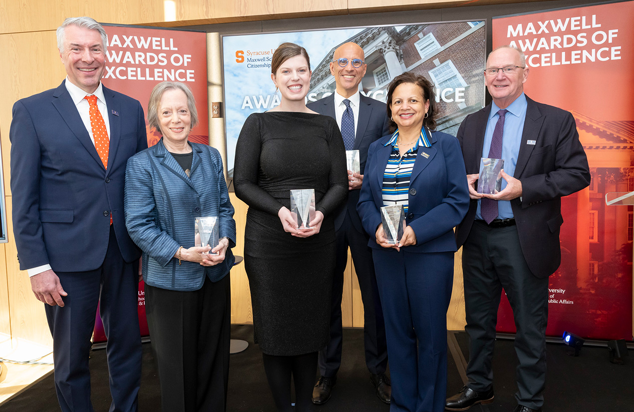 A group of six individuals is standing indoors, each holding a small award. Behind them, banners read "Maxwell Awards of Excellence" and display the Syracuse University logo. They are dressed in formal attire and are smiling at the camera.