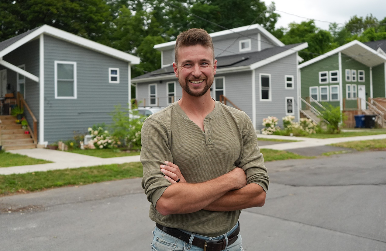 A person stands with arms crossed on a street lined with tiny homes.