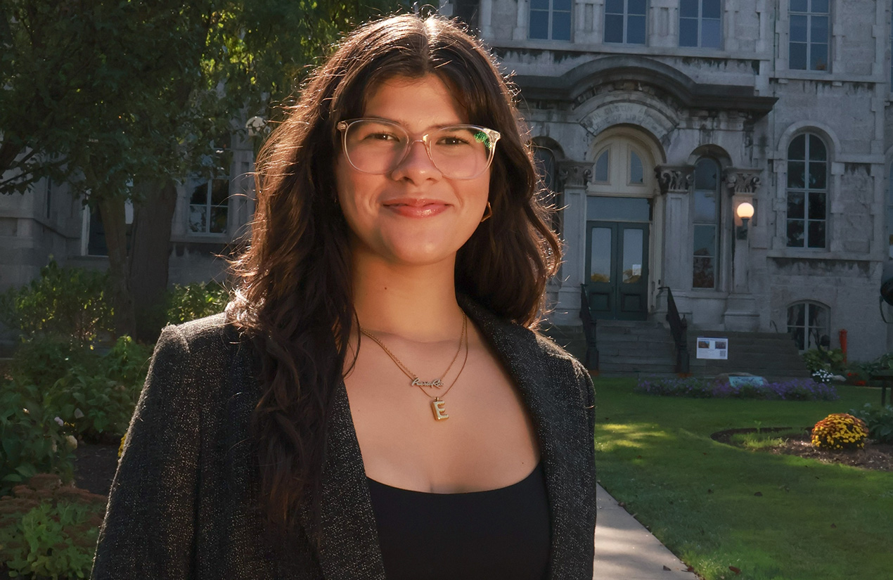 A person with long dark wavy hair and glasses, wearing a black top and gray tweed blazer with a gold necklace, smiling while standing outdoors in front of a stone campus building with a green lawn.