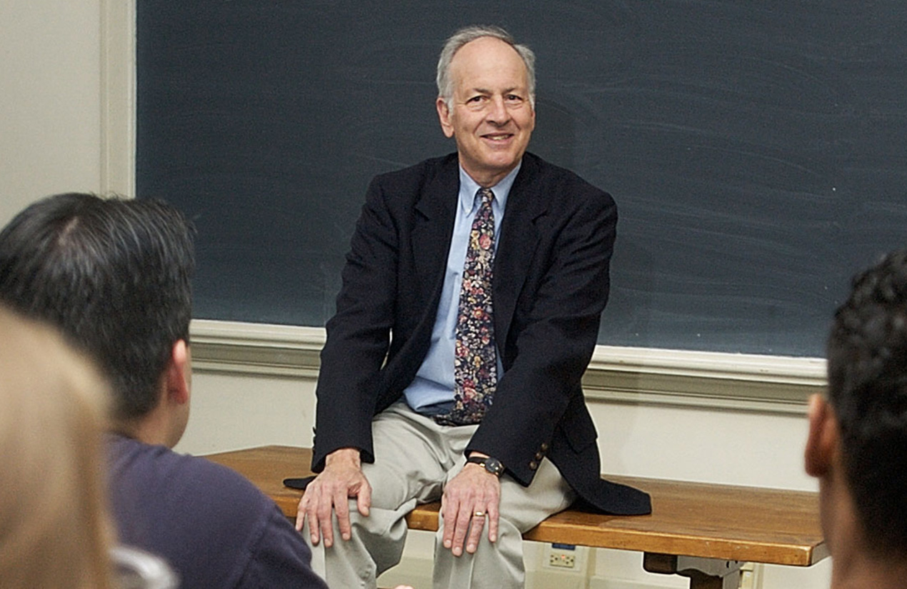 A person sits on a desk at the front of a classroom, smiling and engaged in conversation with students seated in front of him. A chalkboard is visible in the background.