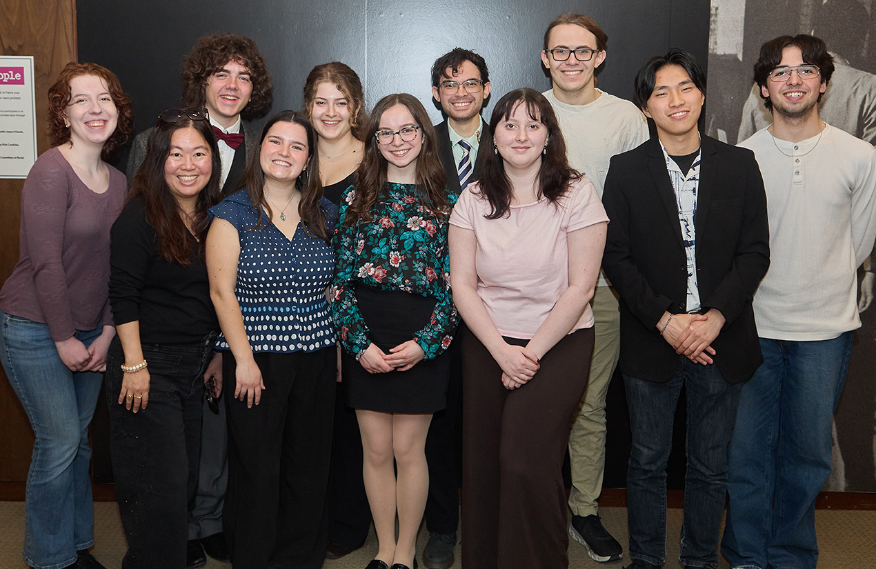 A group of eleven people standing together and smiling for a photo. They are indoors in front of a dark wall, casually dressed.
