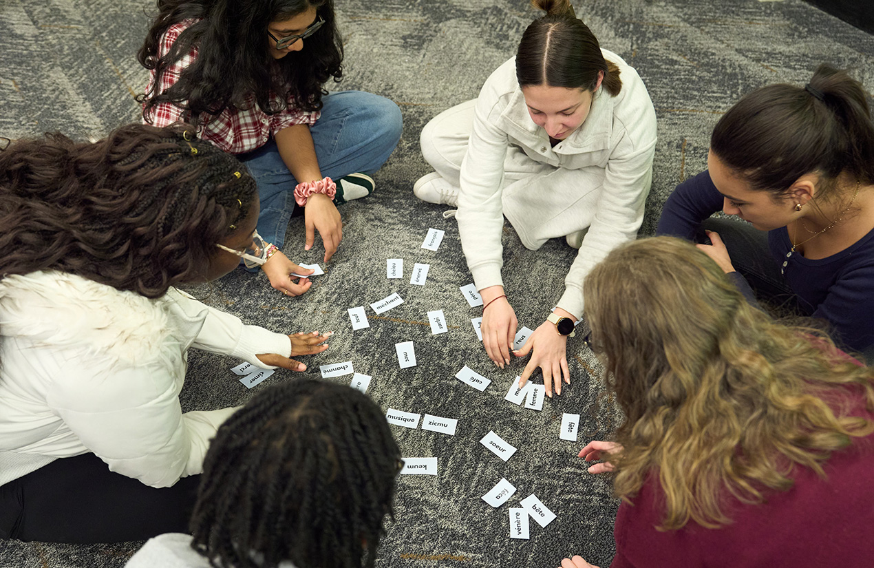 A group of people sitting on a carpeted floor, engaged in a word card activity. The cards are scattered in front of them, and each person is reaching for different cards.