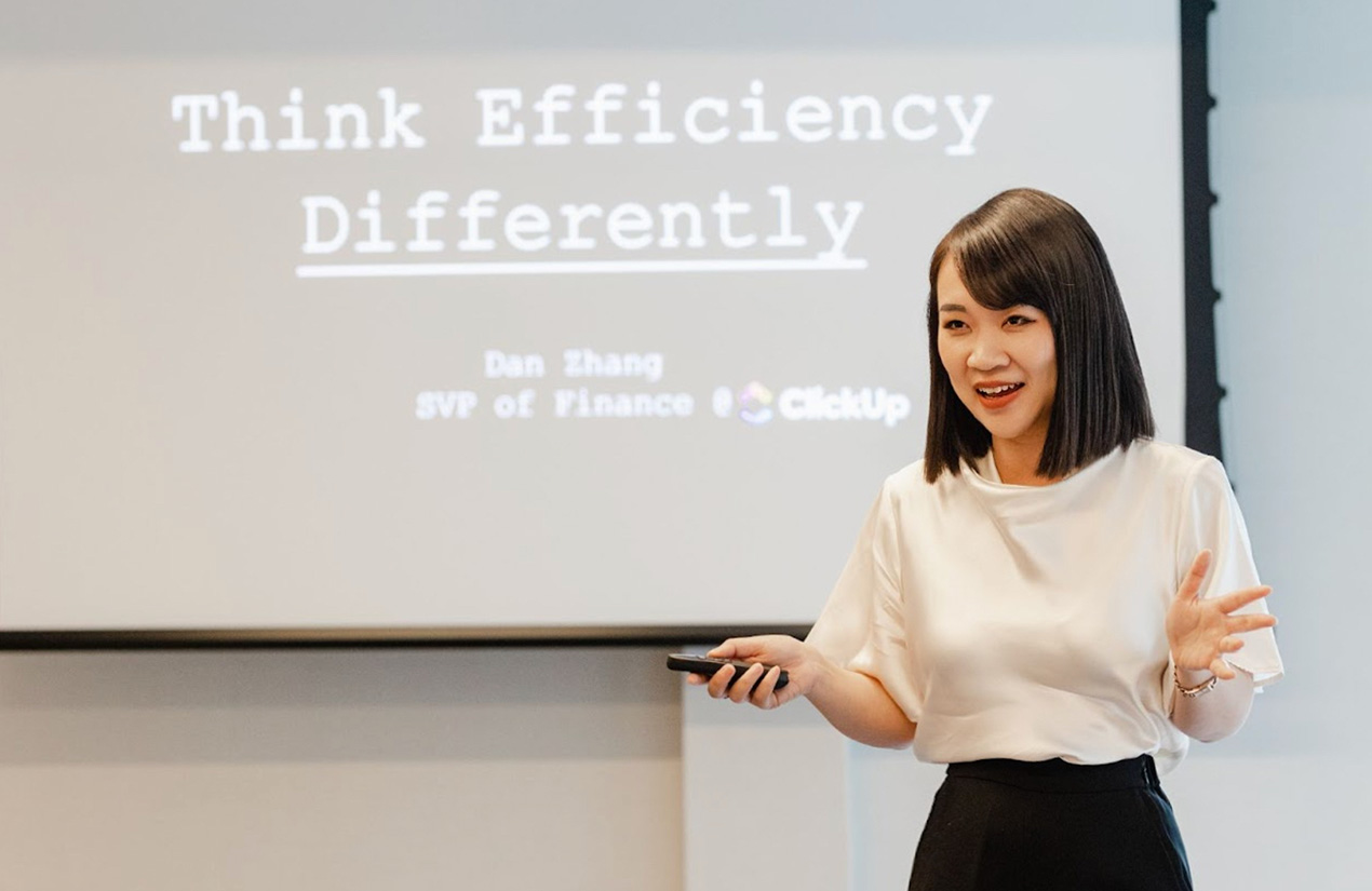 A person stands in front of a presentation screen that displays the phrase "Think Efficiency Differently" along with the text "Dan Zhang, SVP of Finance, ClickUp." The person is holding a remote control and appears to be speaking.