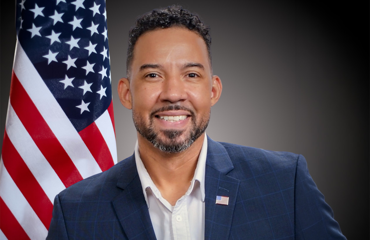 A person wearing a dark suit smiling with an American flag in the background.
