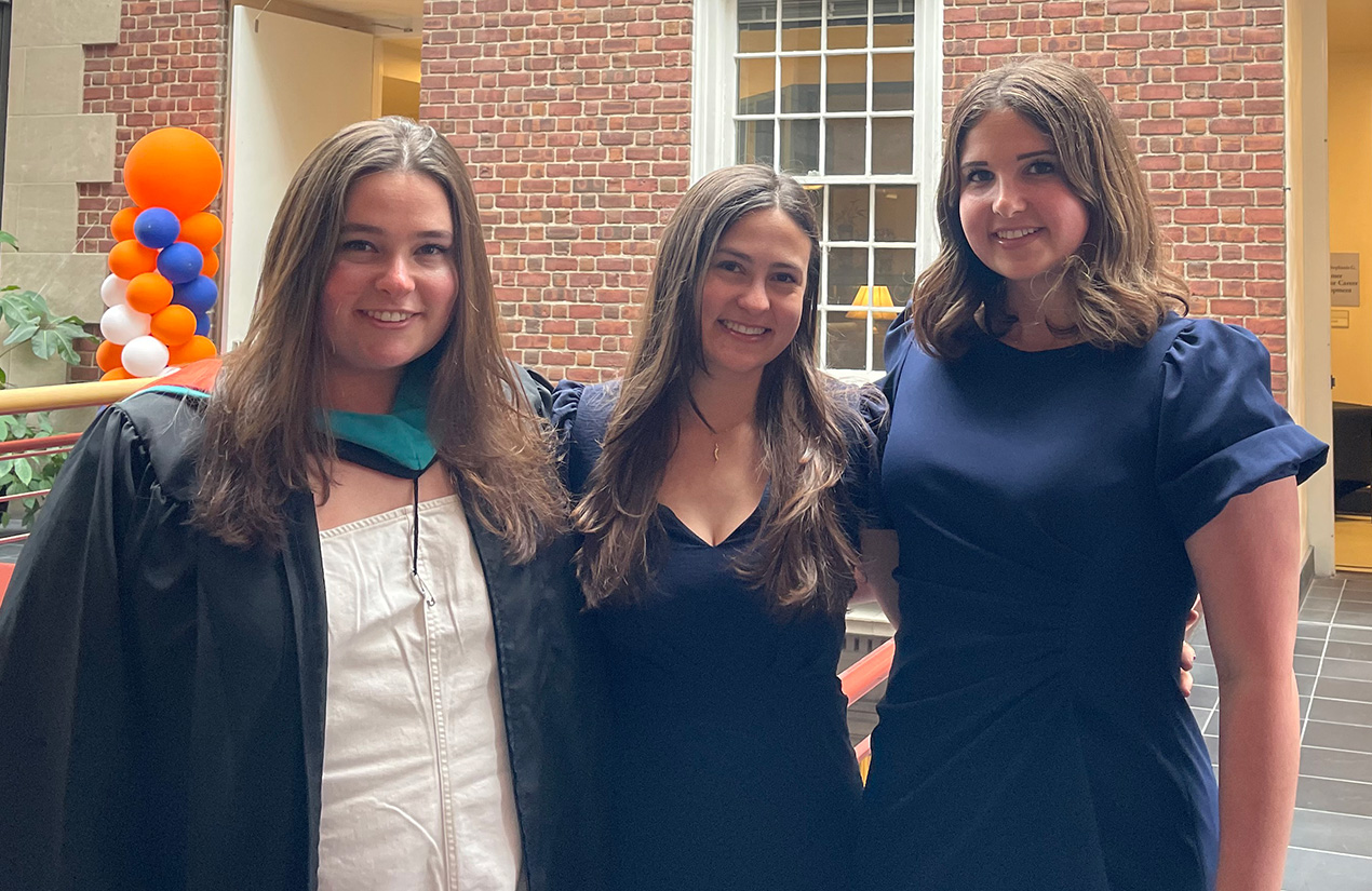 Three people in commencement attire pose together in a brick courtyard.