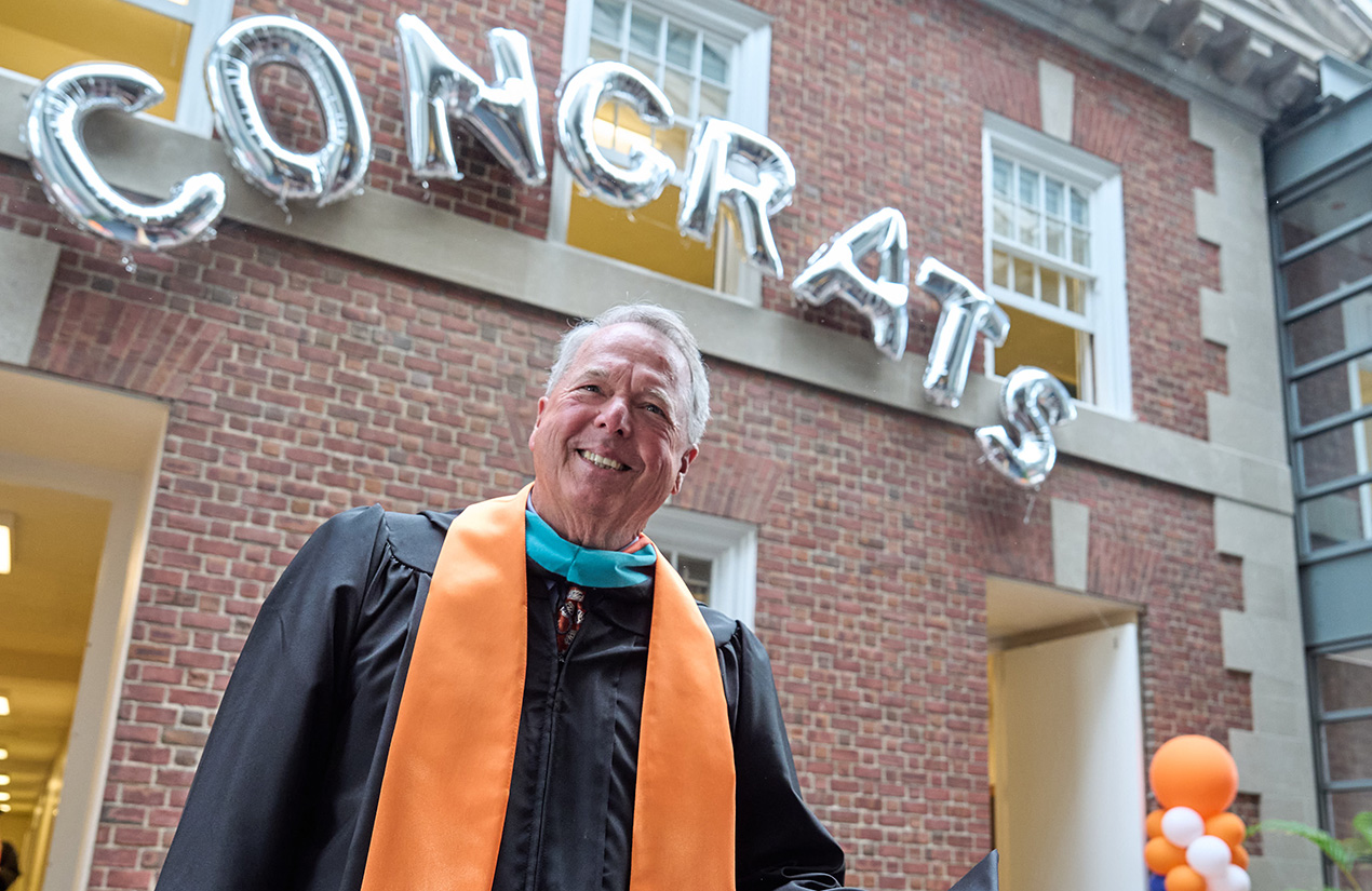 An individual, dressed in academic regalia with an orange stole, stands outside a brick building. Above, silver balloons spell "CONGRATS." Orange and white balloons are visible in the background.