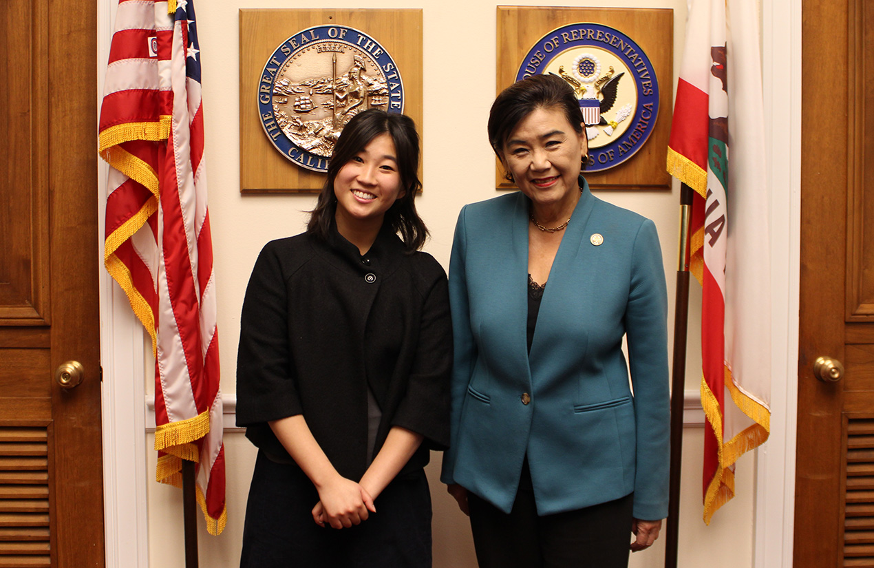 Two people smiling, flanked by an American flag and a California state flag. Behind them are two official seals mounted on the wall, including the Great Seal of the State of California and a U.S. House of Representatives seal.