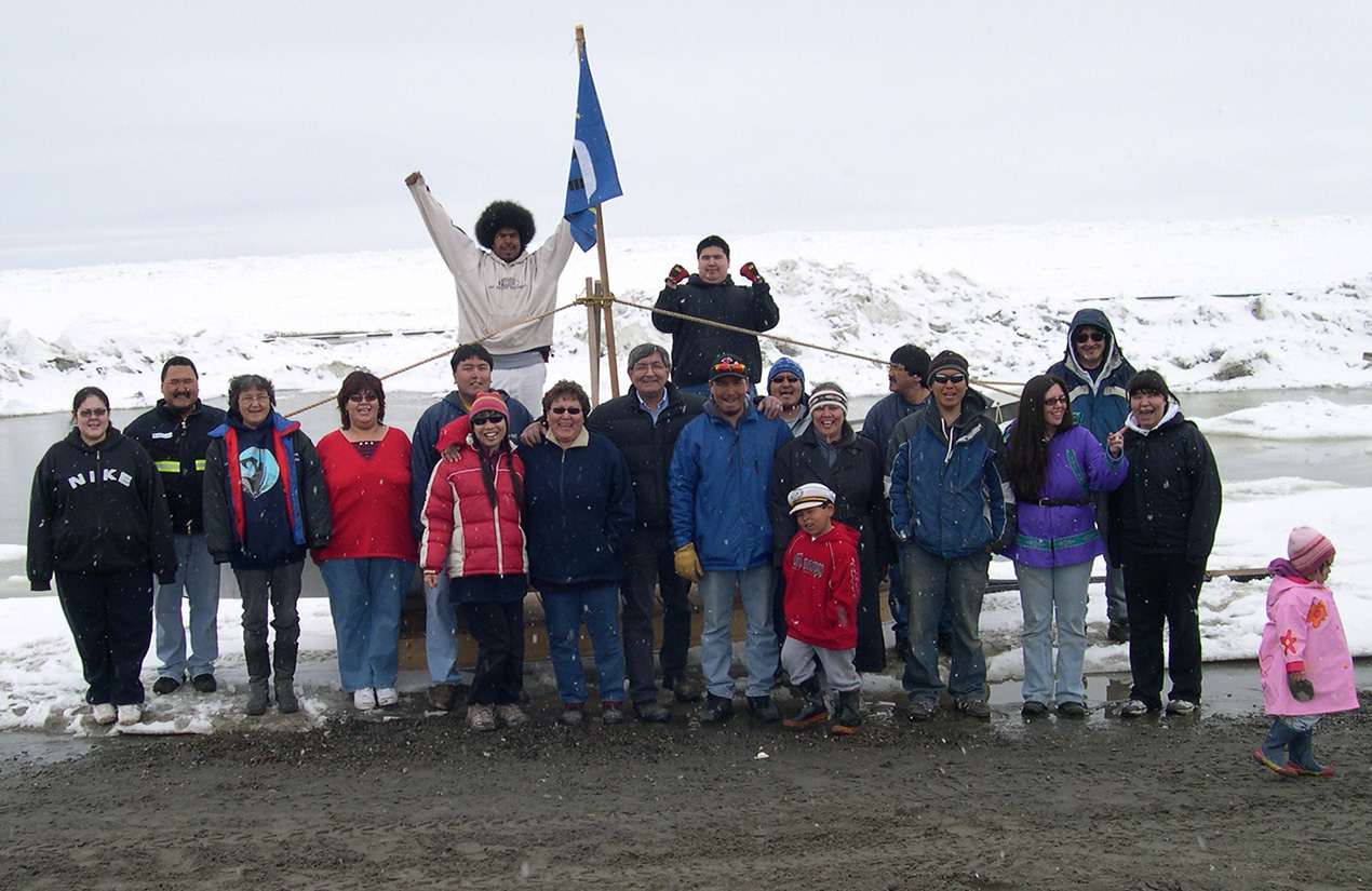 A group of people standing outdoors in a snowy landscape, with some wearing winter clothing. In the background, one person is holding a blue flag with a white emblem. Snow is visible on the ground and in the distance.