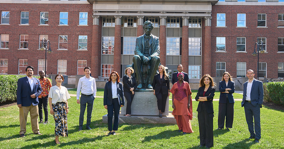 A group of people standing on a lawn, posed around a statue of a seated figure in front of a brick building with tall columns.