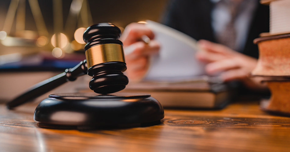 A gavel rests on a sounding block in the foreground. In the blurred background, a person is flipping through a book, with scales of justice visible beside them. The setting suggests a legal or courtroom environment.