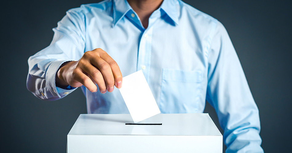 A person in a blue shirt is placing a ballot into a white voting box.