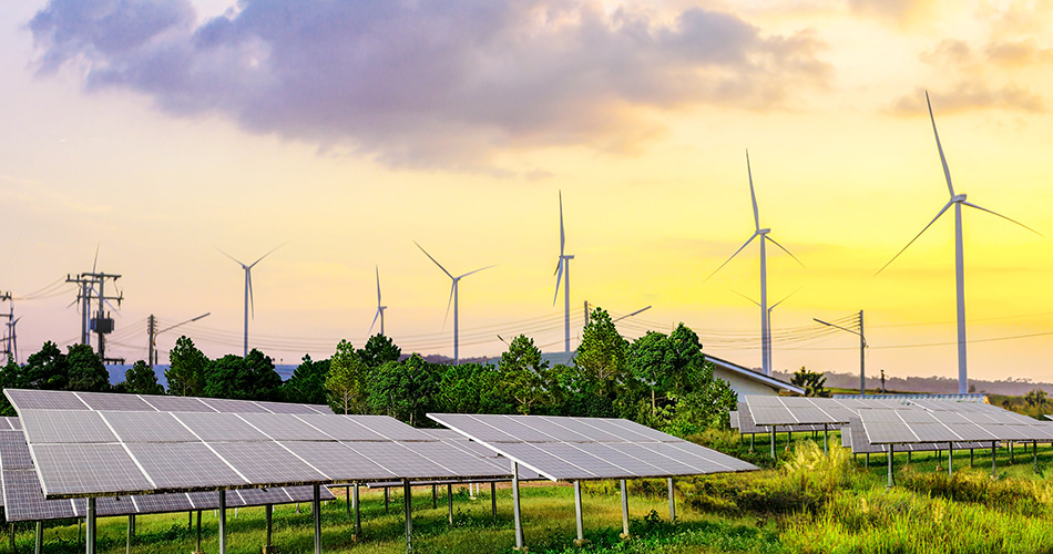 A scenic view of a renewable energy landscape featuring rows of solar panels in the foreground and multiple wind turbines in the background against a sunset sky. Green trees and electric power lines are visible among the renewable energy equipment.