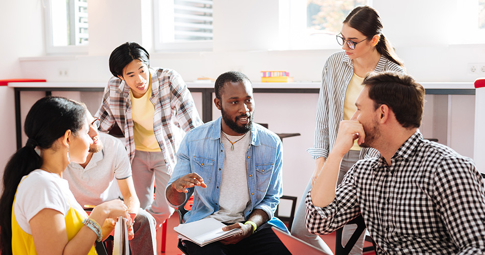 A group of people engaged in discussion while seated in a bright, casual office setting. One person in the center gestures while holding a notebook. They are surrounded by others who are listening attentively.