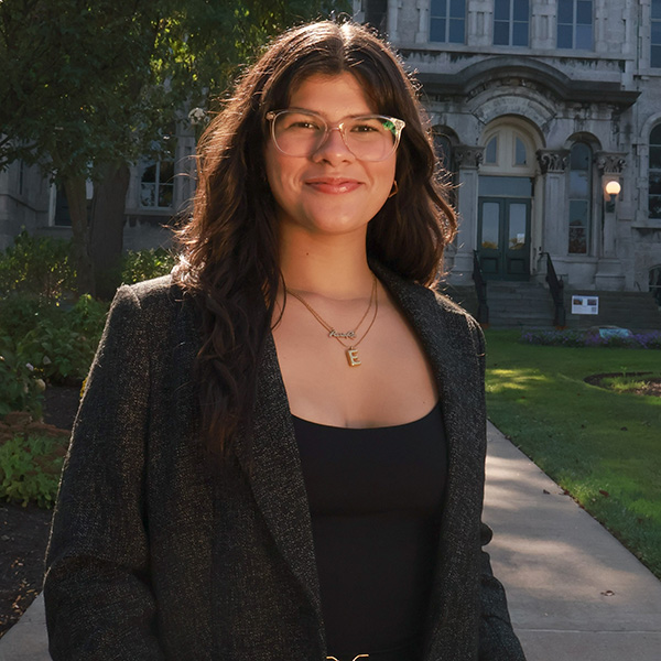 A person with long dark wavy hair and glasses, wearing a black top and gray tweed blazer with a gold necklace, smiling while standing outdoors in front of a stone campus building with a green lawn.