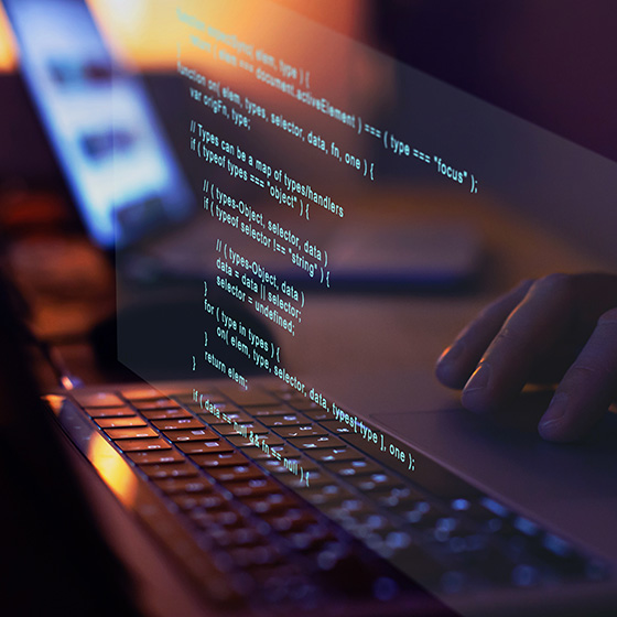 A close-up of hands typing on a laptop keyboard in a dimly lit setting, with lines of glowing blue code projected above the screen.