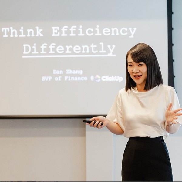 A person speaks at a presentation, gesturing with one hand while holding a clicker, with a projected slide reading "Think Efficiency Differently" visible behind them.