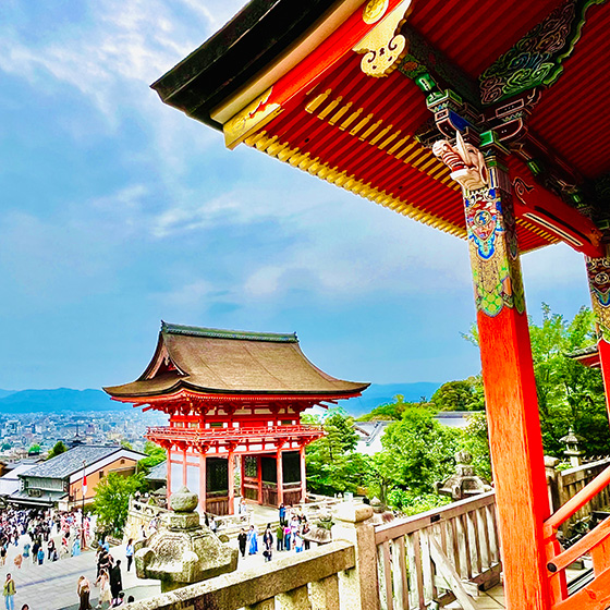 View of Kiyomizu-dera Temple in Kyoto with visitors walking on the temple grounds, cityscape visible in the background.