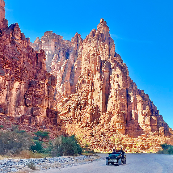 A vehicle drives through a rugged desert landscape dominated by towering red rock formations. Clear blue sky above enhances the vivid colors of the scene.