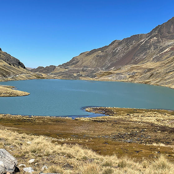 A serene mountain lake under a clear blue sky, surrounded by grassy and rocky terrain with distant rugged hills. The landscape reflects a tranquil natural setting.