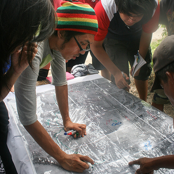 A group of people gather around a large map on a table, discussing and pointing to different areas. One person is writing or drawing on the map with a marker. The scene appears collaborative and focused.