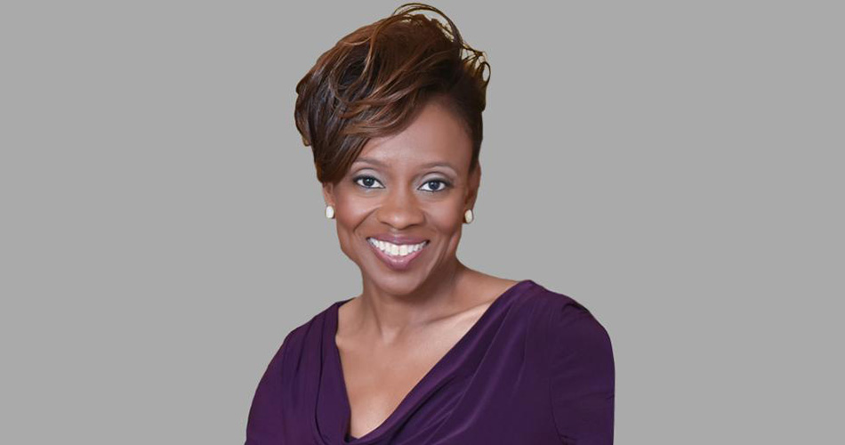 Portrait of a smiling person with short, styled hair, wearing a purple blouse and pearl earrings.