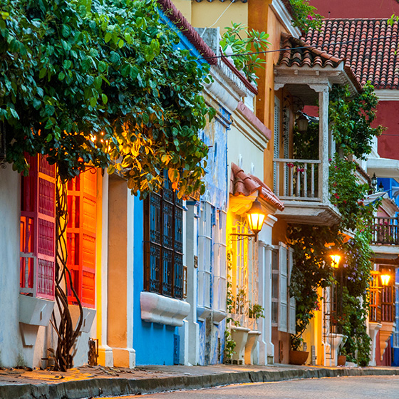 A colorful street lined with vibrantly painted colonial buildings, warm glowing lanterns, and lush trees at dusk in a historic Latin American city.