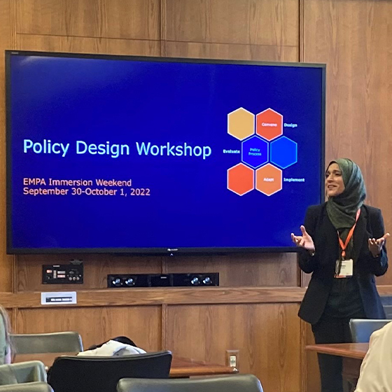 A person is standing in front of a large screen presenting at a Policy Design Workshop during EMPA Immersion Weekend, September 30-October 1, 2022. The screen shows a flowchart with terms: Evaluate, Define, Design, Implement, and Assess.