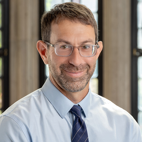 A personsmiling, glasses, short beard, blue shirt and striped tie, near tall windows.